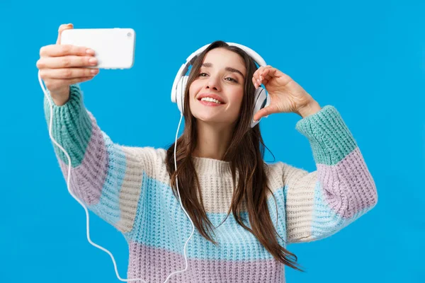 Atractiva, atrevida y elegante mujer morena coqueta en suéter de invierno, tomando selfie usando teléfono inteligente, mostrando el signo del corazón y la cámara del teléfono sonriente, usando auriculares, fondo azul — Foto de Stock