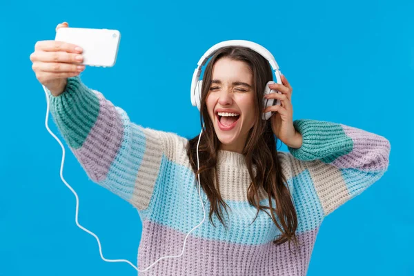 Mujer hermosa alegre, carismática y feliz sonriente en suéter de invierno, disfrutando de música impresionante, tomando selfie como escuchar canción favorita en los auriculares, sosteniendo el teléfono inteligente, fondo azul — Foto de Stock