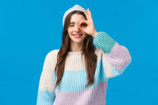 Retrato de cintura arriba alegre hermosa mujer morena en suéter de invierno, sombrero, mostrando bien, buen gesto, signo OK sobre el ojo, sonriendo y guiño en la aprobación, asertivo todo bien, fondo azul — Foto de Stock