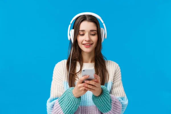 Retrato en la cintura sonriente, mujer guapa feliz con el pelo largo y oscuro, escuchar música en auriculares grandes, sostener el teléfono inteligente, charlar con un amigo, enviar un mensaje, recoger la canción, de pie fondo azul — Foto de Stock