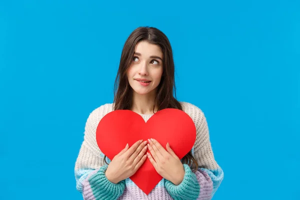 Dreamy lovely and shy, thoughtful attractive woman want give valentines day heart cardboard to express sympathy, confess love and affection, dreamy looking up, biting lip tempting, blue background