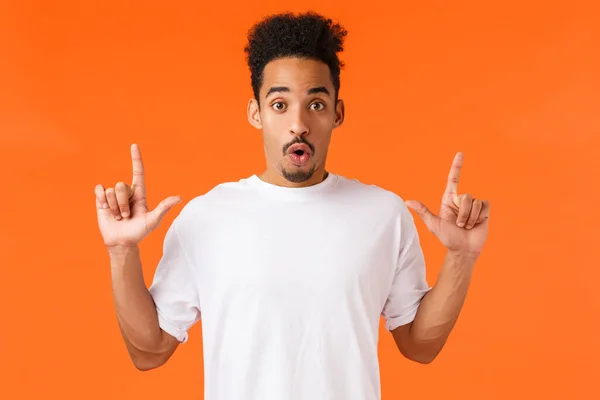 Wow check this out, amazing suggestion. Enthusiastic, intrigued and curious african american male in white t-shirt, pointing fingers up, folding lips gazing impressed, orange background — Stock Photo, Image