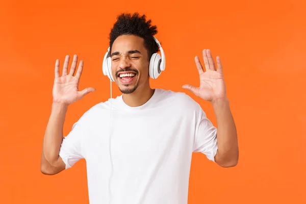 Fantasztikus hangminőség. Gondtalan boldog, mosolygó afro-amerikai férfi fehér pólóban énekel és gesztikulál, mint élvezi a kedvenc zene, zenét hallgatni fülhallgató, csukott szemmel és vigyorgó örömmel — Stock Fotó