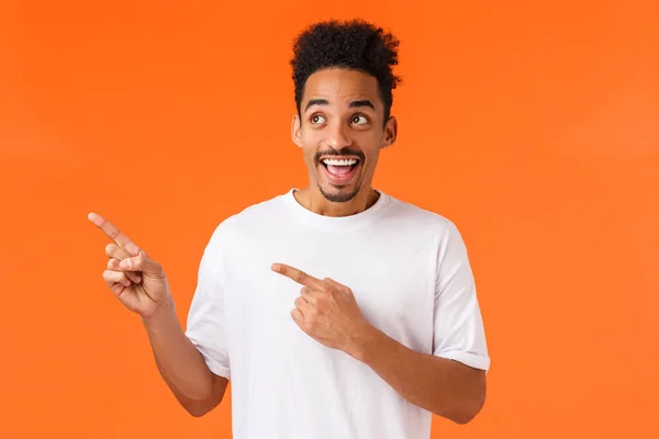 Excited, happy smiling african-american man in white t-shirt, looking wondered and astonished at awesome product advrtisement, pointing upper left corner seeing great discounts, orange background — Stock Photo, Image