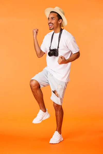 Full-length verticale shot man staan in de luchthaven terminal dragen van korte broek, wit t-shirt, triomfantelijke vuist pomp en kijken linksboven hoek als kijken naar voetbalwedstrijd, winnen, oranje achtergrond — Stockfoto