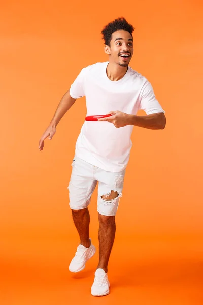 De cuerpo entero vertical tiro feliz, carismático afroamericano deportivo chico como juegos activos al aire libre, lanzando frisbee a amigo y sonriendo, disfrutando de hermoso día de verano soleado, fondo naranja —  Fotos de Stock