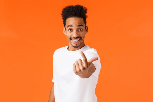 Podekscytowany przyjazny, uśmiechnięty i szczęśliwy afrykański hipster facet amerykański, męski model zwabiający kogoś, wyciągający rękę i machający palcem, aby podejść bliżej, zapraszając osobę, znaleźć kandydata, pomarańczowe tło — Zdjęcie stockowe