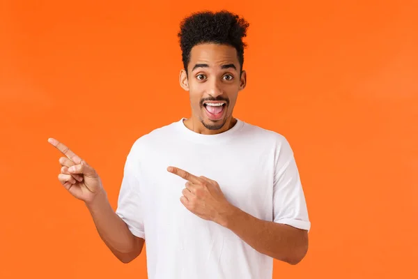 Verás, mira esto. Divertido y emocionado chico hipster afroamericano alegre en camiseta blanca, apuntando esquina superior izquierda y sonriendo, sugerir lugar impresionante, introducir el producto, fondo naranja —  Fotos de Stock