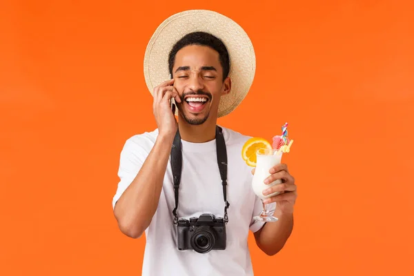 행복 한 웃음이자 카리스마있는, 여름 모자를 쓴 아프리카 계 미국인 남자, 목에 카메라를 매달고, 스마트폰을 하면서 웃고, 휴가중인 누군가에게 전화를 하고, 칵테일을 마시고, 오렌지 배경을 가진 남자. — 스톡 사진