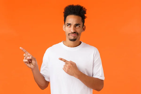 Cético e relutante desapontado homem afro-americano, corte de cabelo afro e bigode, apontando para o canto superior esquerdo descontente, sorrir insatisfeito e julgador, fundo laranja — Fotografia de Stock