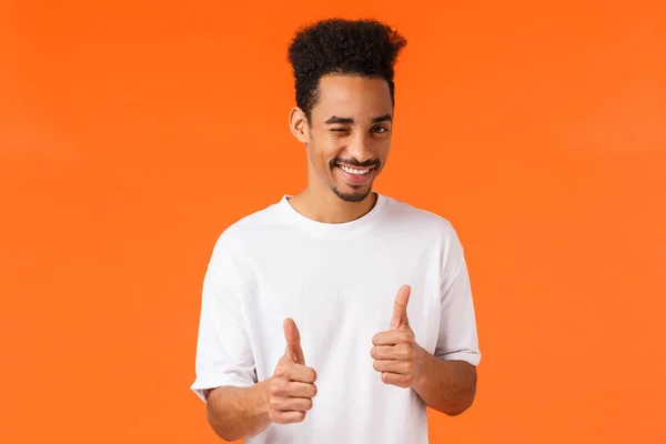 Puedes hacerlo, di que sí. Alegre y confiado joven afroamericano de apoyo en camiseta blanca, guiño y sonrisa, mostrando gesto pulgar hacia arriba, enraizamiento, animar todo lo bueno, buen trabajo —  Fotos de Stock