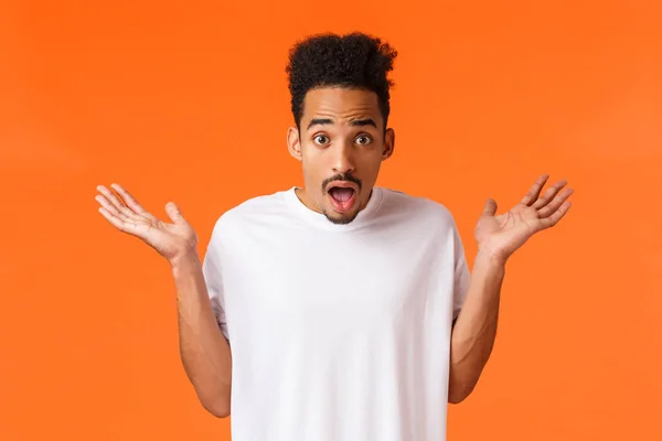 Waist-up portrait amazed, surprised and astonished young hipster modern man in white t-shirt, shrugging raise hands sideways and stare with popped eyes, dropped jaw camera, orange background — Stock Photo, Image