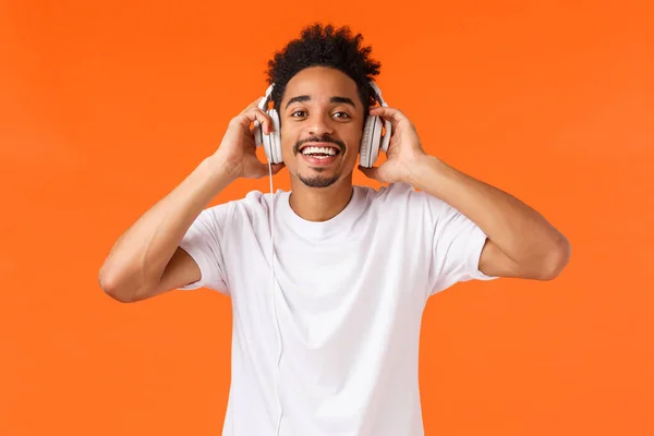Štěstí, technologie a gadgets koncept. Atraktivní šťastný charismatický afroameričan v bílém tričku, poslouchající hudbu ve sluchátkách, usmívající se kamera radostná, jako dárek, oranžové pozadí — Stock fotografie