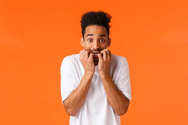 Fear, horror movies, emotions concept. Frightened and insecure, young timid hipster african american male, biting fingernails, staring camera anxious, scared someone know, orange background — Stock Photo, Image