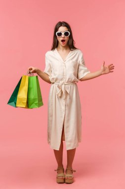 Tam boy dikey portre şaşırmış ve çekici, güneş gözlüklü, elbiseli, çekici bir kadın, elinde çantalar, mağazada birini görmek, nefes nefese, soluk soluğa, pembe arka plan
