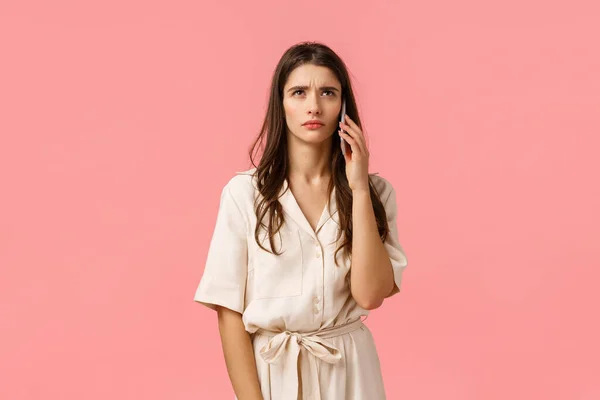 Mujer morena joven seria y estresada escuchando algo extraño, esperando a que alguien coja el teléfono, de pie sobre el fondo rosa en el vestido, sosteniendo el teléfono inteligente cerca de la oreja, fondo rosa —  Fotos de Stock