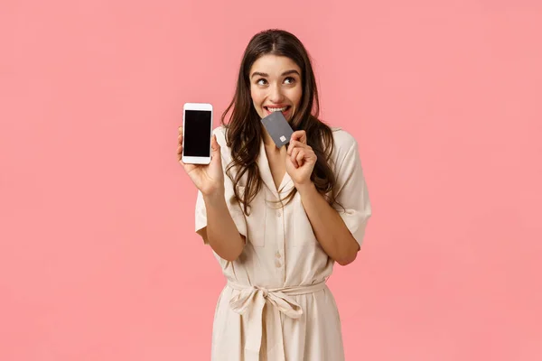 Amused and excited cute girl shoppaholic cant wait for delivery man, order online, biting credit card, holding smartphone, showing mobile screen and gazing sideways dreamy, like spending money — Stock Photo, Image
