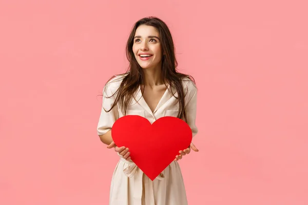 Divertida y despreocupada mujer europea de buen aspecto en vestido, con tarjeta de San Valentín, gran corazón rojo, mirando a la izquierda sorprendida y feliz, sonriente impresionada, de pie fondo rosa alegre —  Fotos de Stock