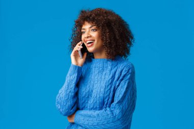 Girl congratulating family with happy holidays, wishing well new year as talking on phone from abroad, holding smartphone speaking, calling friend, looking left and smiling, blue background