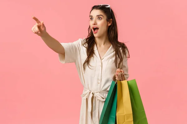 Frau sieht wunderbares Kleid im Geschäft auf Manequin. attraktive, fröhliche junge Frau in Kleid, mit Einkaufstaschen, die zur Seite zeigen und keuchend erstaunt, stehend rosa Hintergrund — Stockfoto