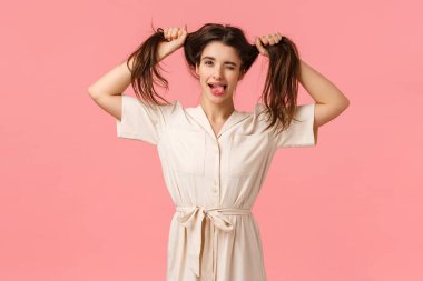 Funny and playful carefree young goofy girl showing tongue and smiling, wink camera happily, pulling hair sideways, making it messy as dont care peoples opinions, standing pink background clipart