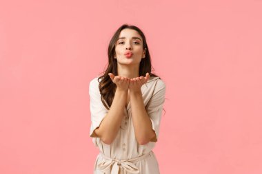 Sevgi ve özen gösteriyorum. Sevimli ve yumuşak Avrupalı esmer, kıvırcık saçlı, bahar elbisesi giyen, elleri katlanmış dudaklara yakın eğilen, havada aşkla öpüşen, pembe arka planda duran