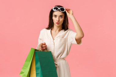Cazibeli beyaz kız eğleniyor, alışveriş torbaları tutuyor, dudaklarını kıvırıyor, eğleniyor, gözlüklerini çıkarıyor, güzel ürünler görüyor, satın almak istiyor, satın alıyor, pembe arka planda duruyor.