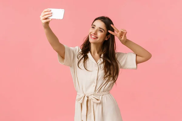 Bekymmersfri känslosam, glad leende brunett kvinna i klänning, hålla smartphone, ta selfie och göra fred tecken, luta huvudet och leende, skicka positiva vibbar till anhängare, stående rosa bakgrund — Stockfoto