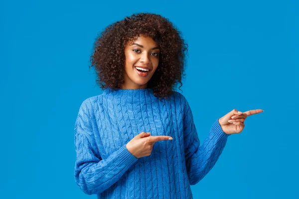 嘿，参观这个网站。穿着冬季毛衣的快乐快乐的非洲裔美国女人，邀请结帐度假价格和特别折扣，指指点点右边，微笑着，蓝色背景 — 图库照片