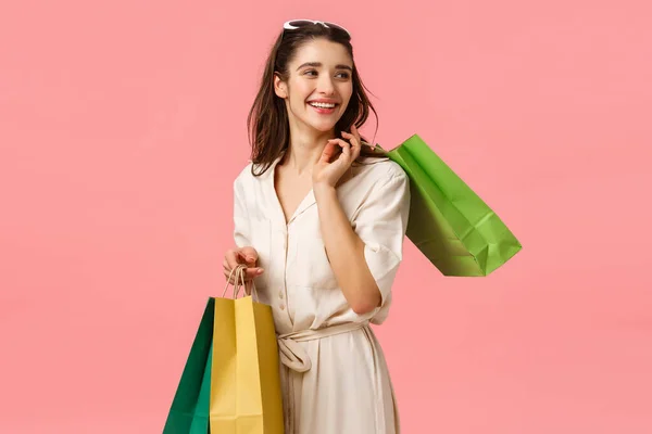 Relajado y despreocupado glamour, chica linda femenina en vestido sosteniendo bolsas de compras y girando a la derecha con hermosa sonrisa feliz, disfrutando de tiendas de moda, de pie fondo rosa —  Fotos de Stock