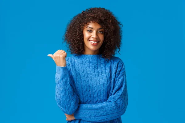Feliz amigable extrovertida, bastante afroamericana hembra en suéter de invierno, compartir enlace, dar consejos, señalar el pulgar a la izquierda para usted check-out anuncio, sonriendo de pie fondo azul — Foto de Stock