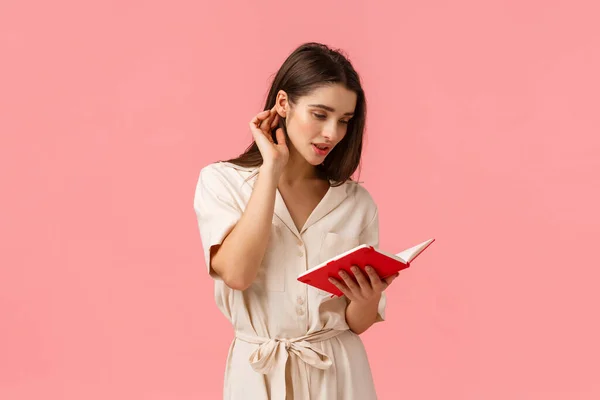 Romantic and tender young alluring woman in dress, put hair strand behind ear and reading notes from red notebook, writing something, studying for exams, standing pink background — Stock Photo, Image