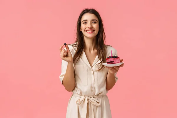 Cumpleaños chica comiendo delicioso pastel, soplando una vela y sonriendo alegremente, sosteniendo arándanos, teniendo fiesta celebrando b-day con amigos, sonriendo de pie en el vestido sobre fondo rosa — Foto de Stock