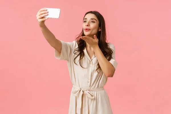 Lovely and tender, alluring brunette female in dress holding smartphone, taking selfie and blowing kiss at mobile camera, stream live video for blog, photographing over pink background — Stock Photo, Image