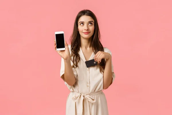 Мечтательная и кокетливая симпатичная молодая женщина в платье, мечтающая о том, чтобы заказать доставку через Интернет, держащая смартфон и кредитную карту, показывающая мобильный экран, стоящий розовый фон — стоковое фото