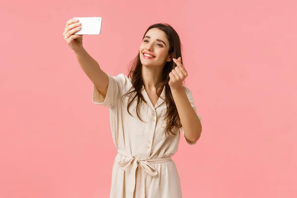 Glada vackra unga europeiska kvinna med filter för att ta söt selfie, fotografering på mobilkamera, leende och visar koreanska hjärta gest på smartphone, stående rosa bakgrund — Stockfoto