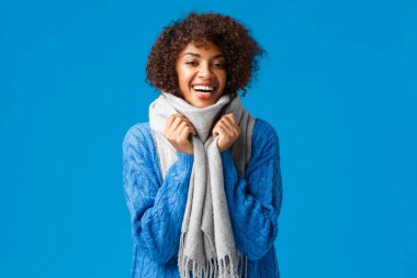 Hoş, karizmatik genç bir kadın kış kıyafetleri aldı, kendini sıcak tutmak için eşarba sardı, gülümsedi ve güldü rahatladı, mavi arka planda dikildi.