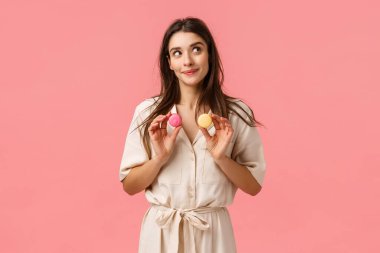 Rüya gibi güzel, tatlı, kadınsı elbiseli kız önce hangi tatlıyı yer, gülümser ve düşünceli görünür, elinde iki badem kurabiyesi, tatlıyı denemek gibi, pembe arka planda neşe içinde durur.