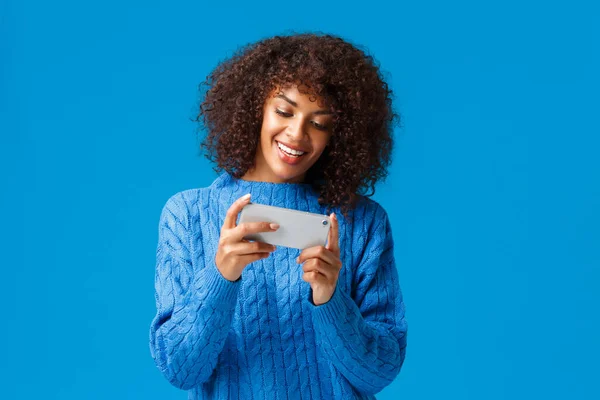Alegre feliz, sonriente joven afroamericana americana jugando juego de teléfonos inteligentes, sosteniendo el teléfono móvil horizontalmente como nivel de carrera que pasa, juego de coches, de pie fondo azul — Foto de Stock