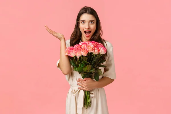 Vaya sorpresa inesperada tan maravillosa. Encantadora chica morena linda y romántica en vestido sobre la pared rosa, recibir hermosas flores, sosteniendo rosas y mirando divertido, levantar la mano asombrado — Foto de Stock