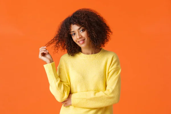 Cintura-up retrato atrevida e coquete boa aparência afro-americana mulher rolando cachos nos dedos inclinar a cabeça e check-out alguém com sorriso atrevido paquera, de pé fundo laranja — Fotografia de Stock