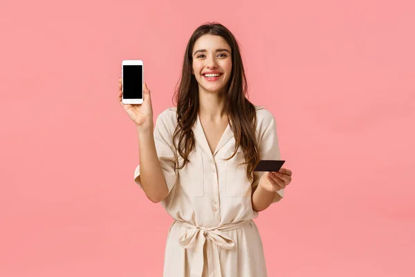 Waist-up portrait happy silly laughing european female making reservetion online, showing cool shopping app on smartphone screen, holding mobile phone and credit card, pink background — Stock Photo, Image