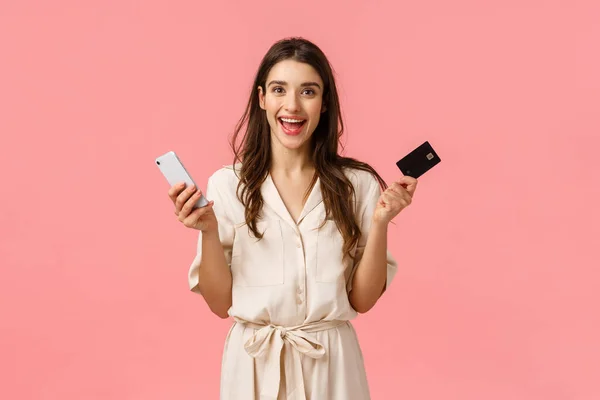 Shopping, online payment, lifestyle concept. Happy alluring girl in dress, holding credit card and smartphone, smiling joyfully as awaiting new product delivery from internet store, pink background — Stock Photo, Image