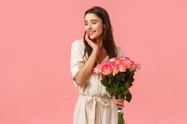 Şefkat, kadınlar ve güzellik konsepti. Çekici, şehvetli bir genç kız, elinde güllerle güzel bir buket çiçek alır, utangaç, sevimli bir gülümsemeyle, pembe arka planda durur.