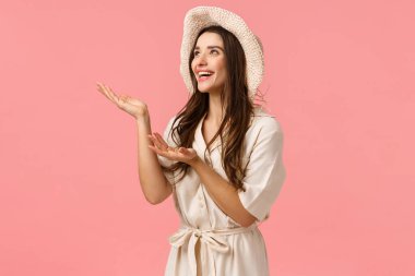 Yaz tatilinde güneşli ve mükemmel havanın tadını çıkaran bir kız. Neşeli, çekici esmer kadın elbise ve şapka ile bir şeyler tanıt, güzel şeylere bak, gülümse, pembe arka plan