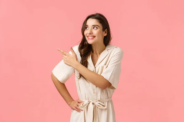 Menina tola antecipando entrega bonito vestido novo para o baile. Mulher morena europeia alegre em vestido ansioso experimentar produto, apontando canto superior esquerdo, sorrindo e rindo, de pé fundo rosa — Fotografia de Stock