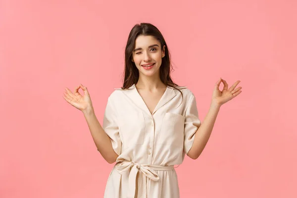 Bleiben Sie ruhig und gehen Sie einkaufen. Fröhlich freche und feminine junge brünette Frau in schönem Kleid, zeigt Zen-Geste, meditiert und zwinkert kokett, stehend rosa Hintergrund — Stockfoto