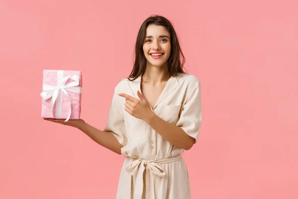 A menina preparou o presente romântico do aniversário. Mulher bonita alegre em vestido bonito segurando caixa de presente e apontando o dedo no presente com encantado, sorriso feliz, de pé fundo rosa — Fotografia de Stock