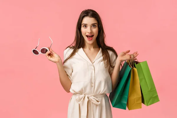 Χαρούμενη πανέμορφη καυκάσια γυναίκα με φόρεμα, κρατώντας τσάντες για ψώνια και γυαλιά ηλίου, χαμογελώντας περίεργος και ενθουσιασμένος, βλέποντας όμορφο ένδυμα σε πάγκους θέλουν να αγοράσουν, στέκεται ροζ φόντο — Φωτογραφία Αρχείου