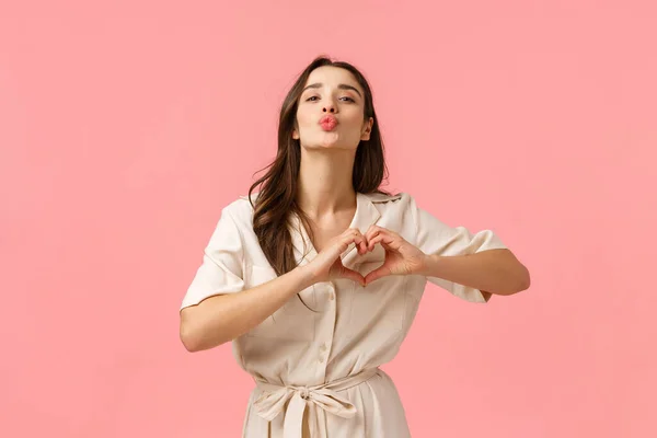 Fashion, beauty and tenderness concept. Charming gorgeous brunette female in cute dress, showing heart love gesture, folding lips for kiss, express romantic feelings, affection, pink background — 스톡 사진